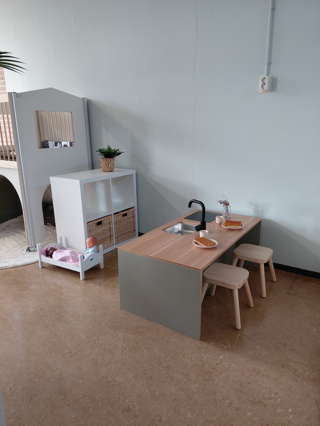 Speelhoek met houten kinderkeuken, krukjes en accessoires in kinderdagverblijf en peuterspeelgroep De Bangert in Andijk, inclusief poppenbed en opbergkast met manden.