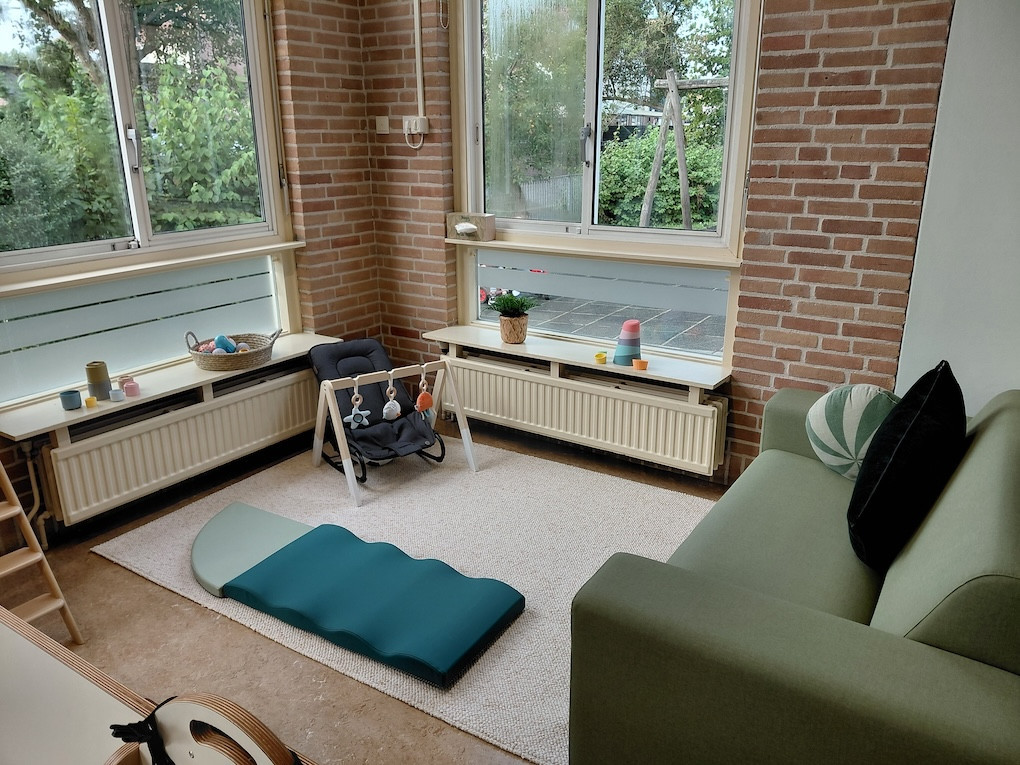 Gezellige speelhoek in kinderdagverblijf en peuterspeelgroep De Bangert in Andijk met groene bank, speelmat, babywipstoel en houten speelgoed bij grote ramen.