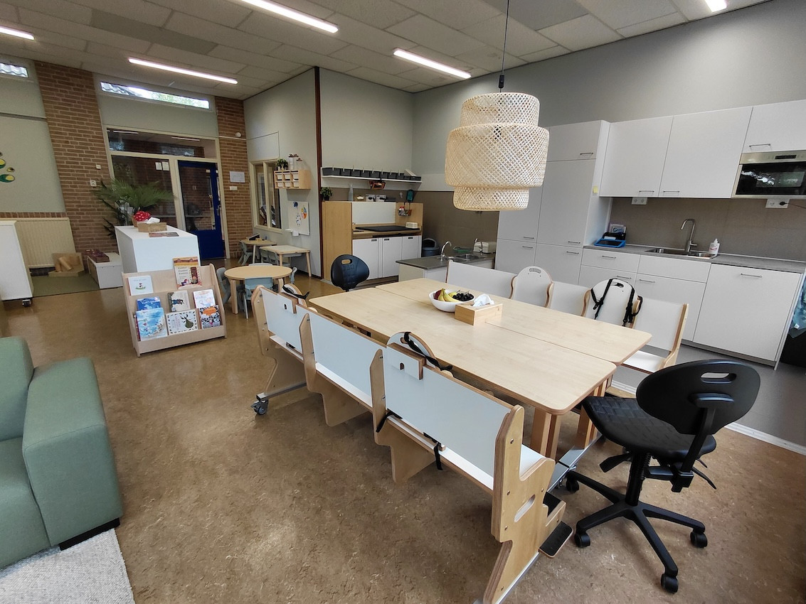 Ruime groepsruimte in kinderdagverblijf en peuterspeelgroep De Bangert in Andijk met grote houten tafel, kinderstoelen, keukenblok en speelhoek.