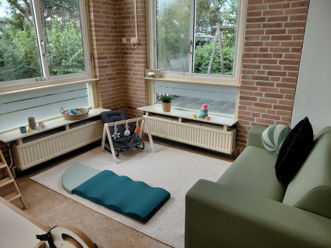 Gezellige speelhoek in kinderdagverblijf en peuterspeelgroep De Bangert in Andijk met groene bank, speelmat, babywipstoel en houten speelgoed bij grote ramen.