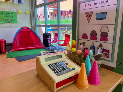 Speelopstelling van een ijswinkel in Peuterspeelgroep De Boevenhoeve. Er is een houten speelgoedkassa, plastic ijsjes en illustraties van verschillende ijssoorten.